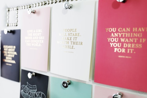 A collection of motivational quotes displayed on different colored cards pinned to a board. Each card has gold lettering featuring different inspirational sayings. The board is organized with silver pins holding the cards in place.