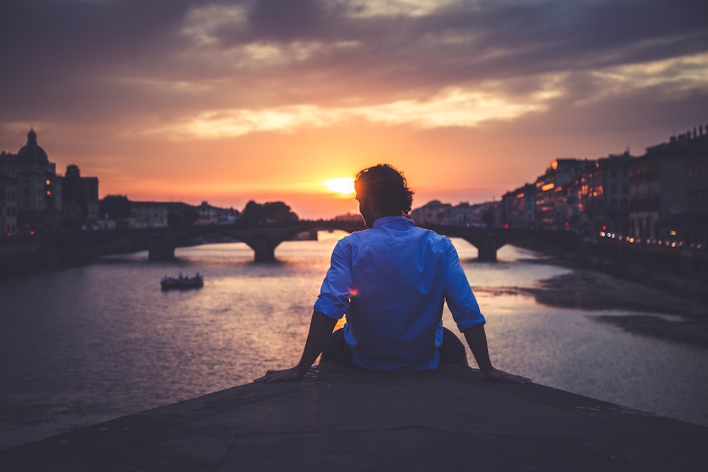 man wearing dress shirt sitting on dock near body of water during golden hour