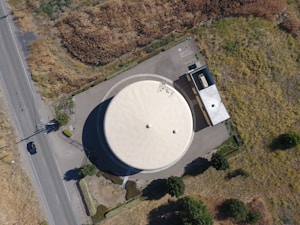 An aerial view of a large, circular water tank with a light-colored roof, surrounded by pavement. Adjacent to the tank is a small rectangular building. The area is bordered by grassy and shrub-covered terrain, with a road running alongside.