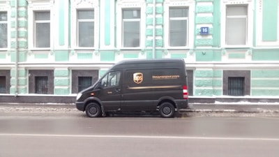 A black delivery van is parked on a street beside a large, ornate building painted in light turquoise with white trim. The van has the UPS logo and some text on its side, and the building features multiple windows and decorative architectural elements.