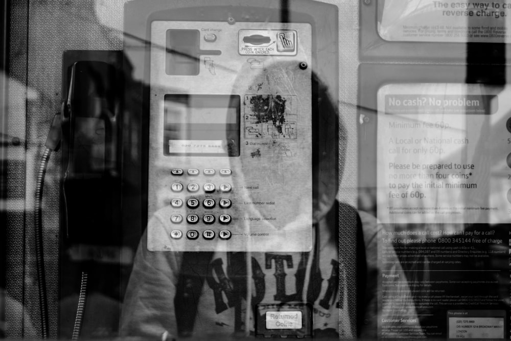 Foto en escala de grises del reflejo de la persona que lleva una sudadera con capucha que lleva una mochila en la pared de vidrio de la cabina telefónica