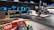 A modern showroom features automotive displays, including an Audi race car in the foreground with various sponsor logos. In the background, there are two dynamic exhibits showcasing the chassis and drivetrain of electric vehicles. A large 'e-tron' sign is prominently displayed, indicative of an electric vehicle focus. Two people are present, taking photos and examining the displays.