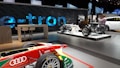 A modern showroom features automotive displays, including an Audi race car in the foreground with various sponsor logos. In the background, there are two dynamic exhibits showcasing the chassis and drivetrain of electric vehicles. A large 'e-tron' sign is prominently displayed, indicative of an electric vehicle focus. Two people are present, taking photos and examining the displays.
