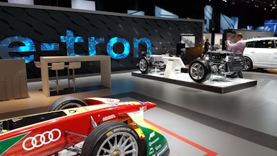 A modern showroom features automotive displays, including an Audi race car in the foreground with various sponsor logos. In the background, there are two dynamic exhibits showcasing the chassis and drivetrain of electric vehicles. A large 'e-tron' sign is prominently displayed, indicative of an electric vehicle focus. Two people are present, taking photos and examining the displays.