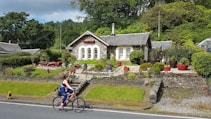 A quaint stone cottage surrounded by a lush garden adorned with colorful flowers and various potted plants. The cottage features arched windows and a sloping roof with a chimney. A woman rides a bicycle along the road in front, and there are trees and greenery in the background, adding to the serene and picturesque setting.
