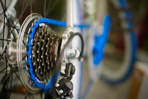 A close-up view of a bicycle gear system with a focus on the cassette and chain. The metallic cogs and blue chain create a contrast against the blurred background of bike parts.