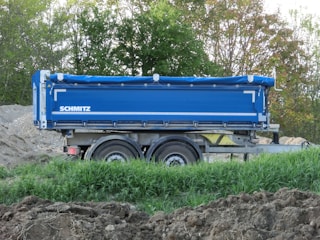A blue trailer with the name 'Schmitz' is parked on a dirt area surrounded by green foliage and trees. The trailer has dual wheels and appears to be designed for transporting goods or materials.
