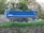 A blue trailer with the name 'Schmitz' is parked on a dirt area surrounded by green foliage and trees. The trailer has dual wheels and appears to be designed for transporting goods or materials.