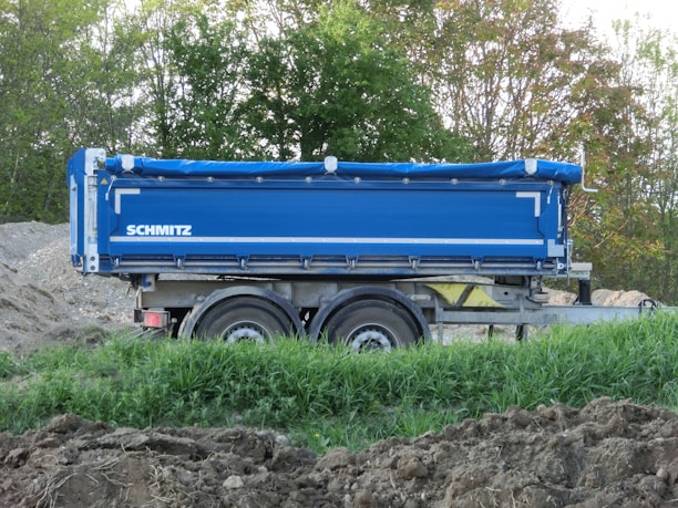 A blue trailer with the name 'Schmitz' is parked on a dirt area surrounded by green foliage and trees. The trailer has dual wheels and appears to be designed for transporting goods or materials.