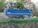 A blue trailer with the name 'Schmitz' is parked on a dirt area surrounded by green foliage and trees. The trailer has dual wheels and appears to be designed for transporting goods or materials.