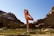 A person is practicing yoga on a rocky landscape with clear blue skies. They are dressed in colorful athletic wear and are balancing on one foot with the other leg extended backward, holding their foot with one hand. The surroundings feature large rock formations and shallow greenish water pooling around the rocks.