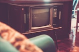A vintage television set with wood paneling stands prominently in a room. The screen is rounded and the knobs and buttons are visible on the side. The surrounding decor is retro, with a teal-colored chair partially in view. The flooring is polished wood, adding to the nostalgic ambiance.