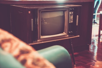 A vintage television set with wood paneling stands prominently in a room. The screen is rounded and the knobs and buttons are visible on the side. The surrounding decor is retro, with a teal-colored chair partially in view. The flooring is polished wood, adding to the nostalgic ambiance.