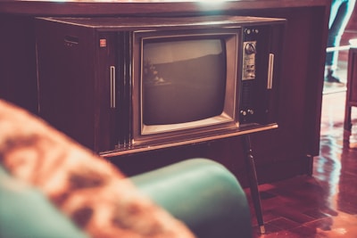 A vintage television set with wood paneling stands prominently in a room. The screen is rounded and the knobs and buttons are visible on the side. The surrounding decor is retro, with a teal-colored chair partially in view. The flooring is polished wood, adding to the nostalgic ambiance.