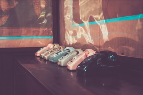 A row of vintage rotary telephones in various pastel colors, including shades of pink, blue, and gray, placed on a dark wooden table against a wall. The setting has a nostalgic, old-fashioned ambiance with warm lighting.