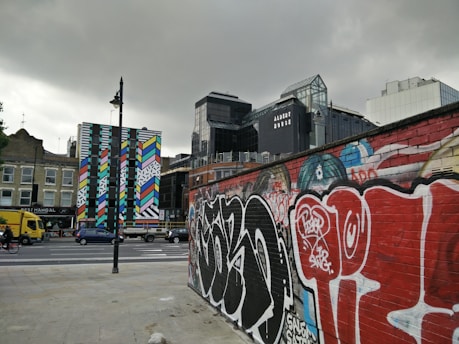 A cityscape featuring a vibrant, colorful mural on a tall, narrow building. The foreground includes a brick wall covered in graffiti. Various modern buildings are visible in the background, including one with the name 'Albert House.' Vehicles, including a yellow truck and a cyclist, are on the road, adding an urban vibe.