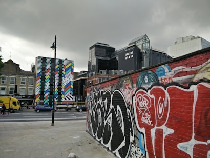 A cityscape featuring a vibrant, colorful mural on a tall, narrow building. The foreground includes a brick wall covered in graffiti. Various modern buildings are visible in the background, including one with the name 'Albert House.' Vehicles, including a yellow truck and a cyclist, are on the road, adding an urban vibe.