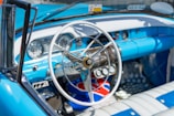 A vintage car interior with a classic steering wheel, chrome accents, and blue and white upholstery. The dashboard features retro gauges and dials, with a small identification card hanging from the rearview mirror.
