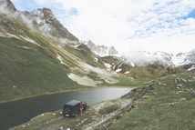 A rugged mountain landscape with a serene lake surrounded by snowy peaks and green hills. A black off-road vehicle is parked near the lake with a person standing beside it. There are patches of snow on the mountains and the sky is partly cloudy.