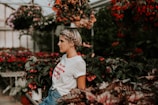 A person with braided hair stands amidst a lush greenhouse filled with various flowering plants, including vibrant red blooms. They wear a white t-shirt with red lettering and blue jeans, and the atmosphere appears serene and contemplative.
