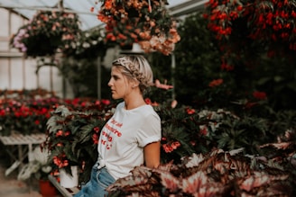 A person with braided hair stands amidst a lush greenhouse filled with various flowering plants, including vibrant red blooms. They wear a white t-shirt with red lettering and blue jeans, and the atmosphere appears serene and contemplative.