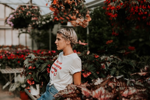 A person with braided hair stands amidst a lush greenhouse filled with various flowering plants, including vibrant red blooms. They wear a white t-shirt with red lettering and blue jeans, and the atmosphere appears serene and contemplative.
