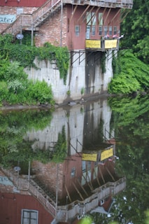 A rustic building made of brick and wood with vines growing along its walls overlooks a calm body of water, reflecting the structure and surrounding greenery. Signs with 'Porcelain' and 'Gifts' are visible, and a wooden staircase leads from the building. The lush greenery envelops the scene, adding a serene and natural element.