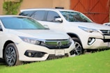 Two white cars are parked close together on a grassy surface. The scene features a Honda sedan in the foreground with a Toyota SUV slightly behind. Both vehicles are clean and polished, reflecting light off their surfaces. There is a building wall and a gate partially visible in the background.