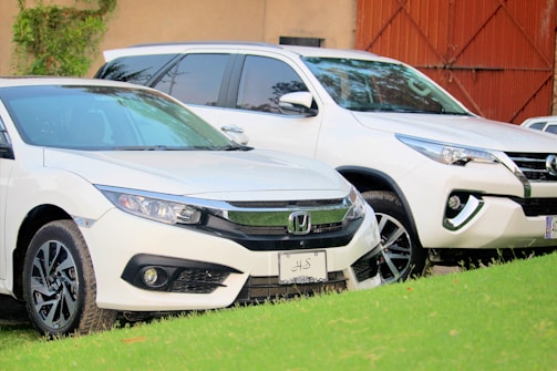 Two white cars are parked close together on a grassy surface. The scene features a Honda sedan in the foreground with a Toyota SUV slightly behind. Both vehicles are clean and polished, reflecting light off their surfaces. There is a building wall and a gate partially visible in the background.