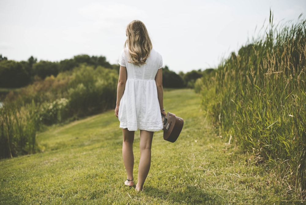 woman holding ukulele while walking on grass field surrounded with tall grasses