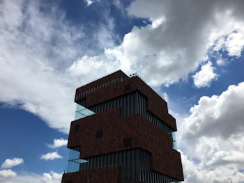A modern, architectural building with stacked, offset layers and large glass windows set against a sky filled with scattered clouds.