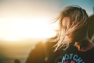 A person with blonde hair is caught in the wind against a backdrop of a bright sunset. The sunlight creates a warm glow, illuminating the strands of hair as they flow across the face. The scene is blurred in the background with hints of an outdoor, possibly coastal setting.