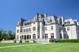 A grand historic mansion with elaborate architectural details stands majestically under a clear blue sky. The mansion features arched windows, ornate carvings, and a steeply pitched roof. Manicured lawns surround the building, and a path leads up to the entrance.