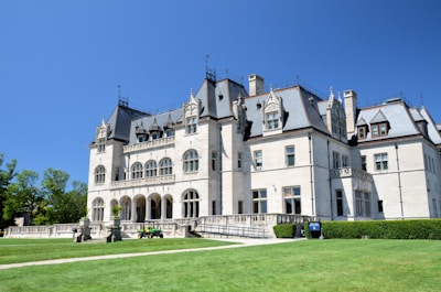 A grand historic mansion with elaborate architectural details stands majestically under a clear blue sky. The mansion features arched windows, ornate carvings, and a steeply pitched roof. Manicured lawns surround the building, and a path leads up to the entrance.