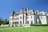 A grand historic mansion with elaborate architectural details stands majestically under a clear blue sky. The mansion features arched windows, ornate carvings, and a steeply pitched roof. Manicured lawns surround the building, and a path leads up to the entrance.