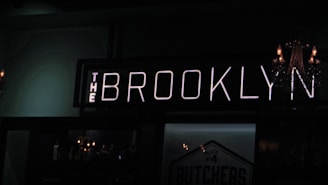 A dimly lit bar with a prominent neon sign reading 'The Brooklyn.' A chandelier with lit candles is partially visible on one side, adding to the subdued ambiance. Bottles are lined up on shelves behind the bar, faintly illuminated by ambient lighting.