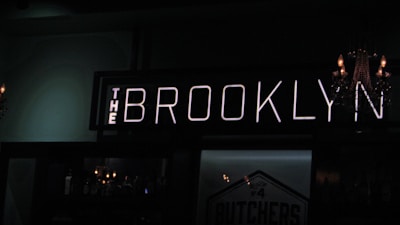 A dimly lit bar with a prominent neon sign reading 'The Brooklyn.' A chandelier with lit candles is partially visible on one side, adding to the subdued ambiance. Bottles are lined up on shelves behind the bar, faintly illuminated by ambient lighting.