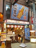 A vibrant shop front features bold signage with Japanese characters and colorful designs. The store sells a variety of goods, possibly food items and condiments, as suggested by the packaging displayed at the entrance. Prominent above the entrance is a large sign depicting a soy sauce bottle with intricate blue patterns surrounding it. Decorative lanterns and banners hang above the open doorway, enhancing the inviting atmosphere.