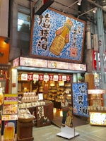 A vibrant shop front features bold signage with Japanese characters and colorful designs. The store sells a variety of goods, possibly food items and condiments, as suggested by the packaging displayed at the entrance. Prominent above the entrance is a large sign depicting a soy sauce bottle with intricate blue patterns surrounding it. Decorative lanterns and banners hang above the open doorway, enhancing the inviting atmosphere.
