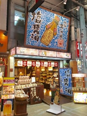 A vibrant shop front features bold signage with Japanese characters and colorful designs. The store sells a variety of goods, possibly food items and condiments, as suggested by the packaging displayed at the entrance. Prominent above the entrance is a large sign depicting a soy sauce bottle with intricate blue patterns surrounding it. Decorative lanterns and banners hang above the open doorway, enhancing the inviting atmosphere.