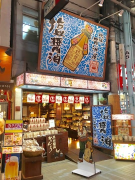 A vibrant shop front features bold signage with Japanese characters and colorful designs. The store sells a variety of goods, possibly food items and condiments, as suggested by the packaging displayed at the entrance. Prominent above the entrance is a large sign depicting a soy sauce bottle with intricate blue patterns surrounding it. Decorative lanterns and banners hang above the open doorway, enhancing the inviting atmosphere.
