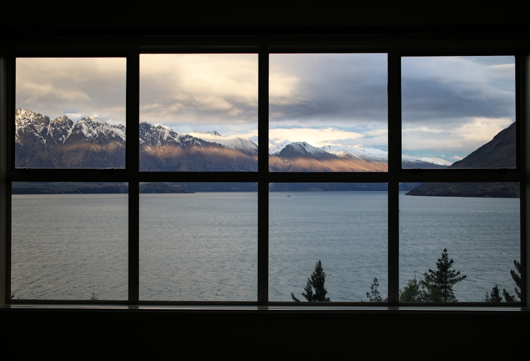 calm body of water near brown mountain under white and gray sky, Taken in Queenstown New Zealand. Window facing Lake Wak
