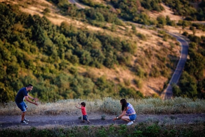 A family scene in a natural setting with a man and a woman encouraging a small child to walk toward them. The landscape features a winding path through a hilly, green area.