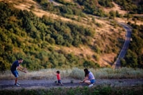 A family scene in a natural setting with a man and a woman encouraging a small child to walk toward them. The landscape features a winding path through a hilly, green area.