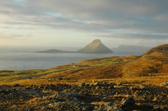 A serene landscape featuring a vast expanse of rocky terrain leading to the ocean. In the distance, a distinctive cone-shaped island is visible under a partly cloudy sky, with the warm light of the setting sun casting golden hues on the landscape.