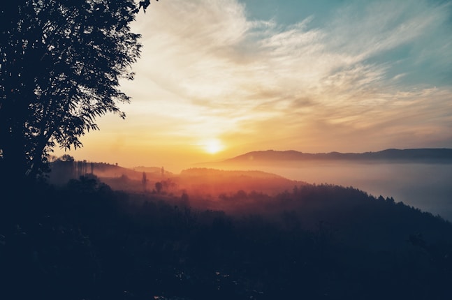 A beautiful sunrise over a misty mountain landscape with a vibrant sky and silhouettes of trees in the foreground.