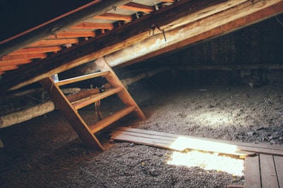 An attic with a wooden ladder leaning against an inclined ceiling. Sunlight streams through gaps, illuminating parts of the space. The floor appears to be covered with gravel or small stones.