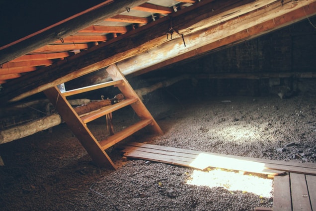 An attic with a wooden ladder leaning against an inclined ceiling. Sunlight streams through gaps, illuminating parts of the space. The floor appears to be covered with gravel or small stones.