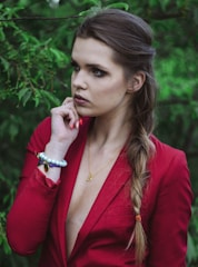 A woman with a serious expression is surrounded by lush green foliage. She is wearing a red blazer with a deep neckline and has a long braid draped over her shoulder. Her hand is raised to her chin, displaying a bracelet with large beads and a butterfly charm. A delicate necklace rests around her neck.