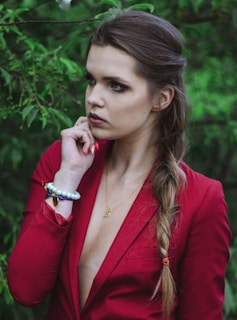 A woman with a serious expression is surrounded by lush green foliage. She is wearing a red blazer with a deep neckline and has a long braid draped over her shoulder. Her hand is raised to her chin, displaying a bracelet with large beads and a butterfly charm. A delicate necklace rests around her neck.
